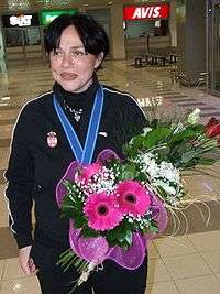 A laughing middle-aged woman with a flower bouquet and a medal around her neck, wearing a Serbia national team tracksuit.