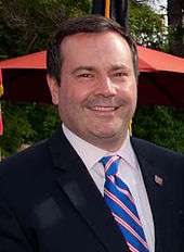A photograph of a man outside wearing a black suit, a white dress shirt, and a blue striped tie, looking at the viewer and smiling
