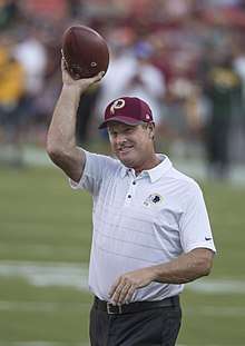 Color photograph of smiling white man (Jay Gruden), wearing a white sport shirt and holding a football.