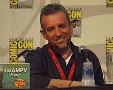 A Caucasian man in his forties, seated at a conference, with a microphone in front of him. He has a pleasant square face, deep-set eyes, greyish hair and a brown beard with clean-shaved cheeks and upper-lip. He is casually dressed, relaxed and smiling. Square signs are posted on the wall behind him, bearing the name COMIC-CON in big bright yellow letters around a drawn eye and eyebrow.