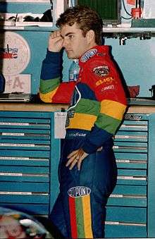 A man his late twenties wearing rainbow colored racing overalls. He has his left hand on his left hip and his right hand leaning against a solid surface.