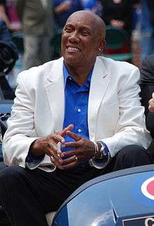 A smiling, dark-skinned man wearing a white suit jacket, a blue button-down shirt and dark pants