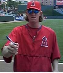 A white man wearing a red baseball cap and warmup, both with a red "A" topped by a halo. The man is holding out his right hand, which is holding a pen, and wearing sunglasses.