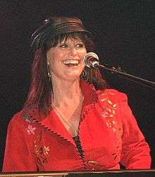 A woman with long brown hair wearing a black cap and a red shirt, sitting at a microphone