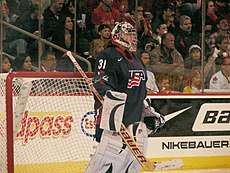 A female ice hockey goaltender is on the ice.  She is standing straight and looking forward.  Her jersey is blue and features a stylized USA.
