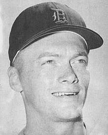 A black-and-white headshot of a smiling man wearing a baseball cap with an Old English "D" on the face