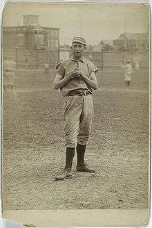 A sepia-toned image of a young man in an old-style white baseball uniform and cap standing on a grass field holding a baseball in front of his chest with both hands