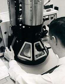 A black-and-white photograph of a young man examining a large microscope. The man has short, dark hair, is wearing a white shirt and a white lab coat and is holding a smoking pipe in his mouth. The microscope has a black conical base with three trapezoidal windows and a silver cylindrical body.