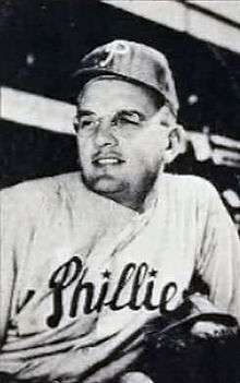 A black-and-white image of a man in a white pinstriped baseball uniform with "Phillies" across the chest, a dark baseball cap with a white "P" on the front, and round glasses