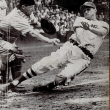 A black-and-white photo of a man wearing a baseball uniform sliding feet-first into home plate