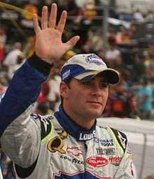 A man in his early thirties waving to the crowd with his right hand and is wearing white and blue racing overalls along with a similarly colored baseball cap