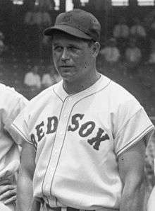 A man in a white baseball jersey reading "RED SOX" across the chest and a dark baseball cap looks toward the left of the image