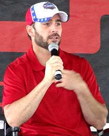 A man in his mid thirties speaking into a microphone. He is wearing a red and white baseball cap and a red t-shirt.