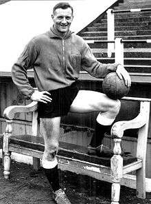 Posed monochrome photo of white man in football kit standing with one foot up on a bench.