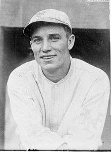 A black-and-white image of a smiling man wearing a pinstriped crownless baseball cap and a white baseball jersey
