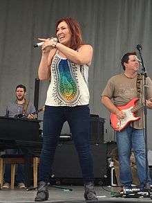 A woman with long brown hair wearing a white top with a multi-coloured pattern and blue jeands, singing into a microphone in front of two men playing guitars