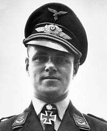 A black and white photo of the head and shoulders of a young man. He wears a peaked cap and military uniform with an Iron Cross displayed at the front of his shirt collar.