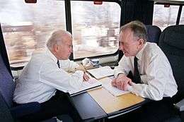 Two men speaking to each other in a compartment on a train. Sitting in blue seats with paperwork spread out on tables in front of them, the man on the left has white hair and the man on the right has gray hair. Both wear white dress shirts, black pants and ties.