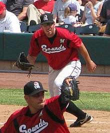 A man with a black cap, red baseball jersey with "Sounds" written across the chest in white, and white pants leans forward at first base preparing to catch a ball