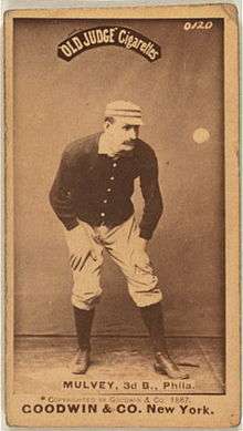 A sepia-toned baseball-card image of a man wearing an old-style black baseball sweater, white pants, and a pillbox hat