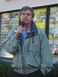 A gray-haired man wearing a green jacket speaks into a microphone outside in front of a building, with campaign signs that read "Joe Sestak" and "Obama-Biden" in the background.