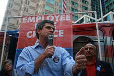 A gray-haired man wearing a blue shirt and black campaign button speaks into a microphone at an outside political rally. A man and woman stand behind him, and a banner reading "Employee Free Choice" can be seen in the background.
