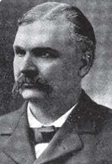 A man with graying hair and a dark mustache wearing a black jacket and tie and white shirt