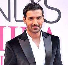 A smiling John Abraham, in a white shirt and black coat