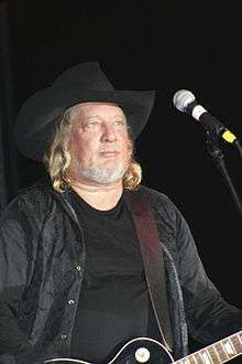 A man with long blond hair wearing a black cowboy hat and black clothing, playing a guitar
