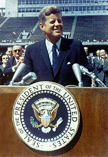 Kennedy, in a blue suit and tie, speaks at a wooden podium bearing the seal of the President of the United States. Vice President Lyndon Johnson and other dignitaries stand behind him.