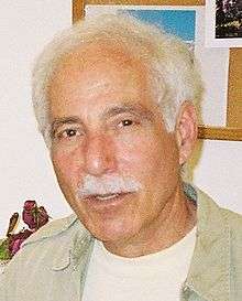 Casual portrait photograph of male, age appearing to be in 50's (chronologically 70's), with brown eyes, brown eyebrows, white hair and mustache, wearing white t-shirt and khaki shirt.