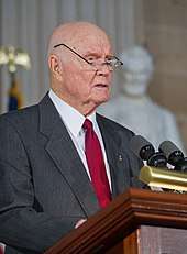 An older John Glenn speaking at a podium, with his glasses perched high above his ears so he can read with them