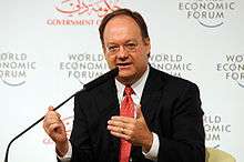A balding white male seated behind a microphone wearing a typical suit and red tie. Behind him is a white wall with logos for the World Economic Forum.