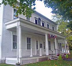 A white house with porch and fluted columns decorated in red, white and blue bunting viewed from its left with some tree branches visible at the sides. A small sign near the front steps says "John Kane House".