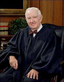 Man in shirt and bow-tie and judge's robes seated in a chair