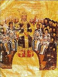 Sitting elderly man in black and gold robes, wearing a golden crown and holding scepter in the center. Behind and around him, arranged in a semicircle, are seated bearded men, some in white and others in black dress. Bearded heads of other men with tubular and triangular hats are visible behind them.
