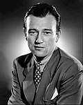 Black and white photo of John Wayne in 1940—a white man with a broad forehead, dark straight hair and dark eyes, wearing an elegant suit, around 34 years of age.