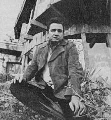 A black-haired man sitting on the grass in front of a wooden building