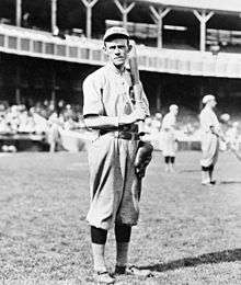 A black-and-white photograph of a man in an old-style white baseball uniform holding a baseball bat over his left shoulder; he is standing on a grass field in a stadium