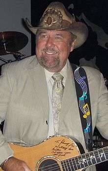 A heavy-set middle-aged man with a beard, wearing a cowboy hat, grey jacket and tie, playing a guitar