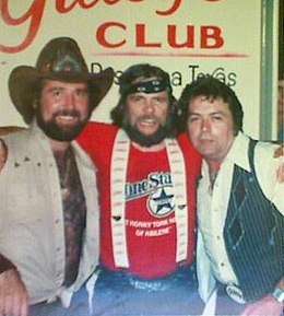 A group of three men with their arms round each other.  The man on the left has a beard and is wearing a cowboy hat and a light-coloured vest.  The one in the center has long hair and a beard and is wearing a bandana tied around his forehead, a red T-shirt and suspenders.  The one on the right is wearing a white shirt and a dark vest.