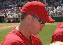 A man in profile, wearing a red baseball jersey and red baseball cap