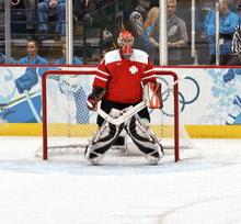 An ice hockey goaltender on ice in front of a net. He is wearing a red and white sweater, and black and grey pads.