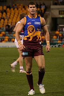 A Caucasian man in a maroon, blue and gold Australian rules football jersey walks towards the camera on grass