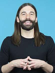 Image of Jonathan Van Ness in front of a blue background. He has long, straight brown hair and a mustache. He's smiling and is holding his hands in front of his chest.