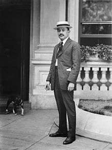 A black and white portrait of a formally dressed young man with a short, black mustache wearing a light-colored hat, white shirt, a light colored suit, dark tie and dark shoes. The man is outside a building where a dog is coming out.