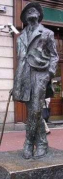 Bronze statue of Joyce standing in a coat and broadbrimmed hat. His head is cocked looking up, his left leg is crossed over his right, his right hand holds a cane, and his left is in his pants pocket, with the left part of his coat tucked back.