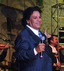 A man in a blue suit holding a microphone. He is smiling and extending his arm towards the back of a stage, where two men are playing stringed instruments.