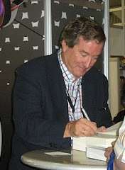 A white man sits in front of a desk, while signing a book and smiling.