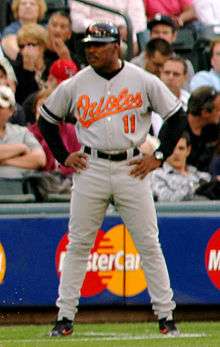 A dark-skinned mustachioed man wearing a gray baseball uniform with orange script across the chest and standing with arms akimbo